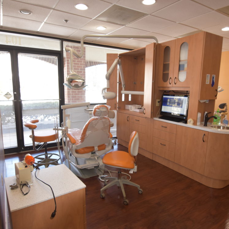 This is an interior view of a dental office, featuring a modern dental chair and equipment on the right side of the image.