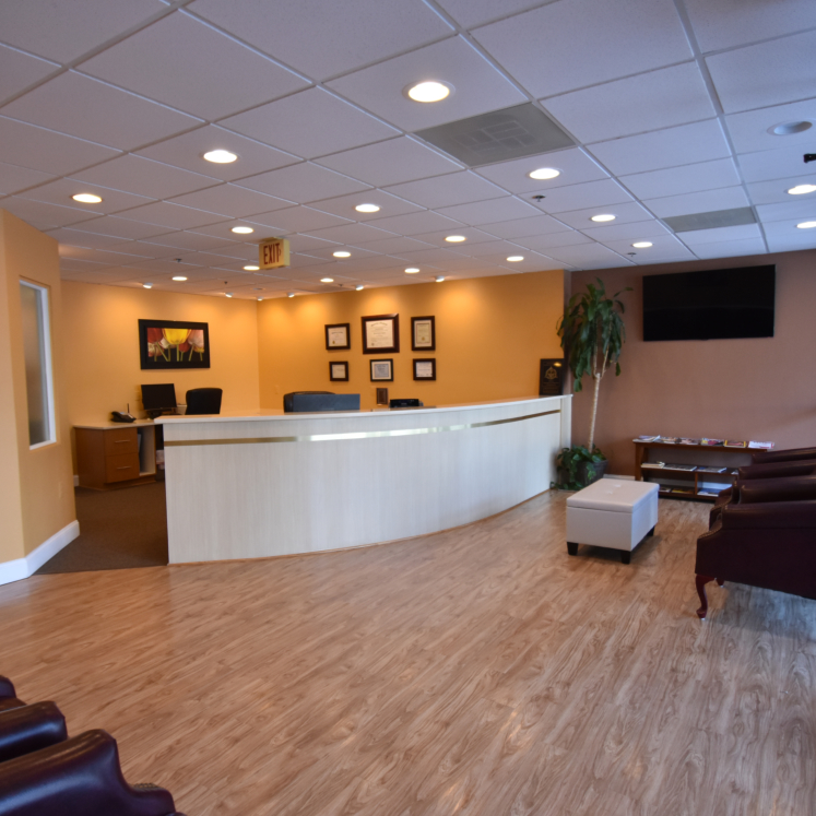 The image shows an interior view of a modern office reception area with a clean, bright design, featuring a reception desk, seating area, and framed artwork on the wall.