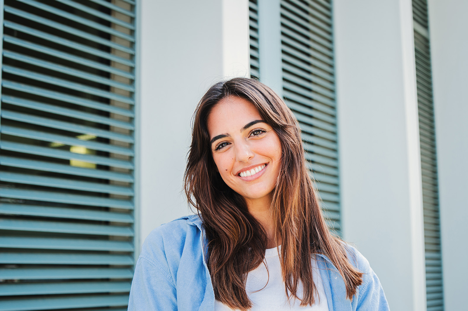 The image shows a person with long hair, smiling at the camera, standing in front of a building with vertical slats.