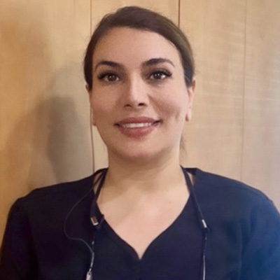 A woman standing against a wall, smiling and wearing a black shirt with a name tag.