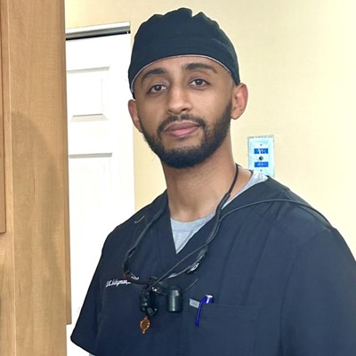 A man in a black surgical gown and cap, standing with his hands slightly apart from his body, wearing glasses and a lanyard around his neck.