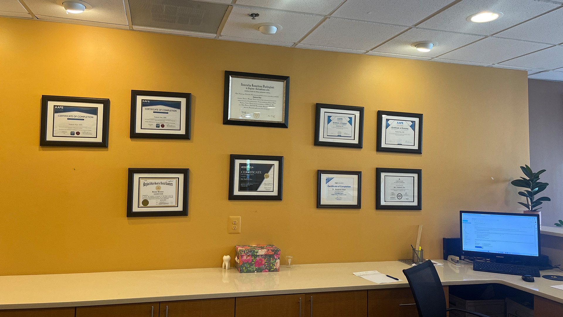 The image displays a wall with multiple framed certificates, set against a yellow background.