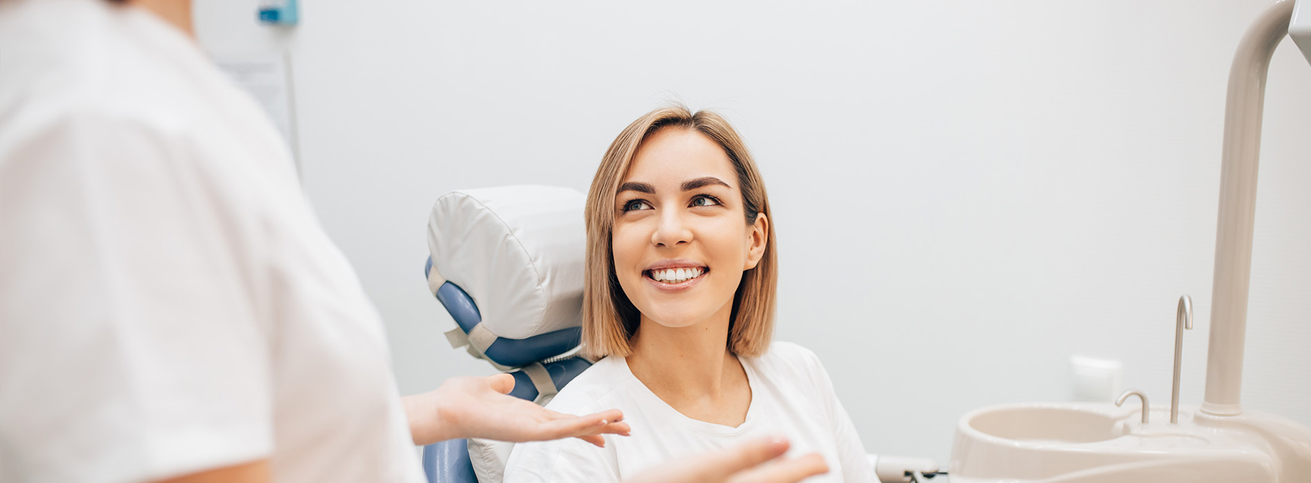 Woman in white shirt sitting in dental chair with smiling dental hygienist standing behind her.