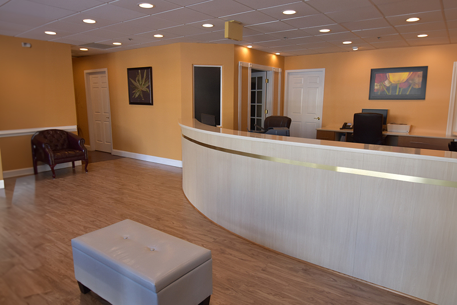 The image shows an interior view of a modern office reception area with a light-colored floor, a curved counter with a decorative top, and a seating area with a bench and a stool.