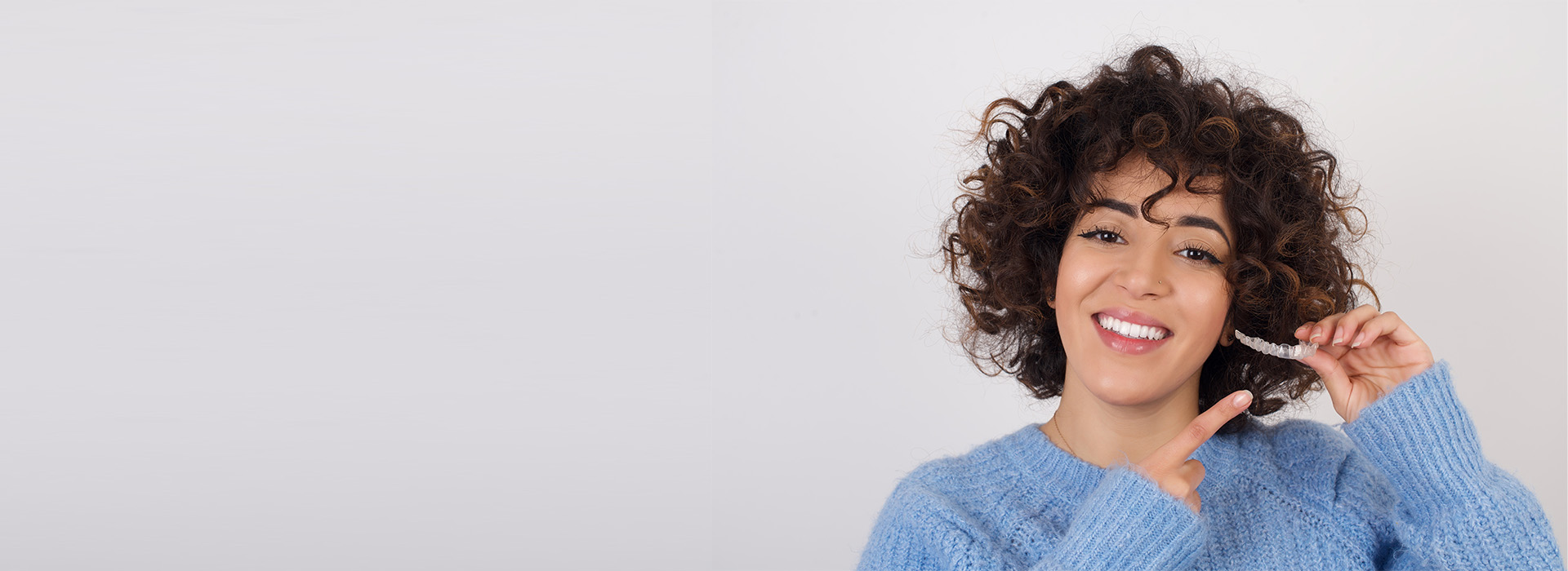 The image shows a person with curly hair smiling at the camera, wearing a blue sweater and holding a smartphone. They are against a plain background.