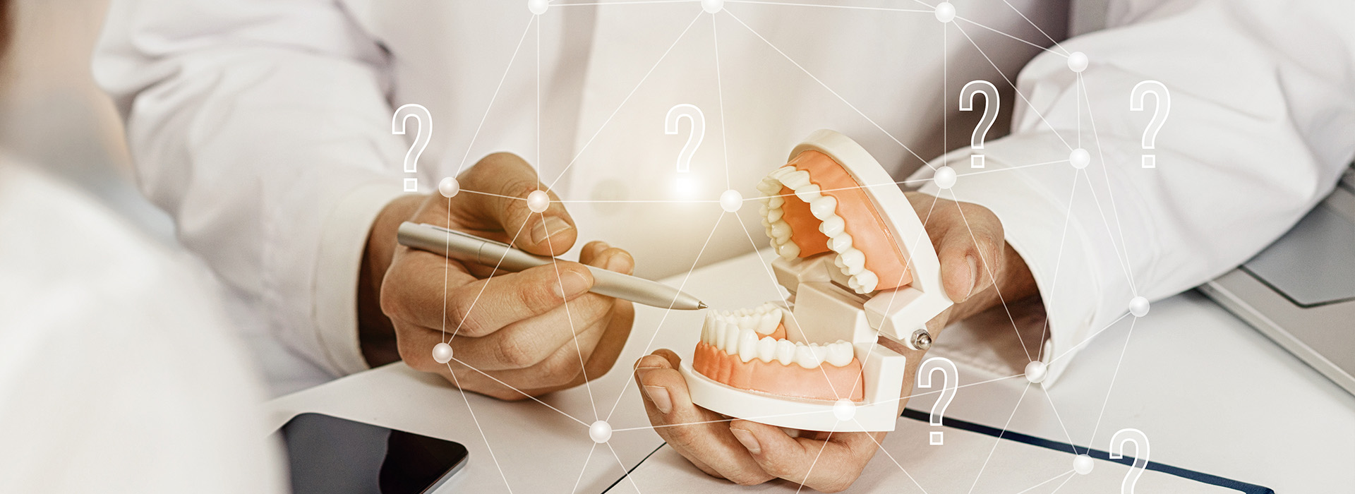 The image shows a person s hand holding a model of a human mouth with teeth, in front of a digital display that includes icons and numbers. The setting appears to be a dental or medical office, as suggested by the presence of what looks like a dental implant on the desk and the professional attire of the individual. The image has an overlaid watermark or logo at the bottom right corner.