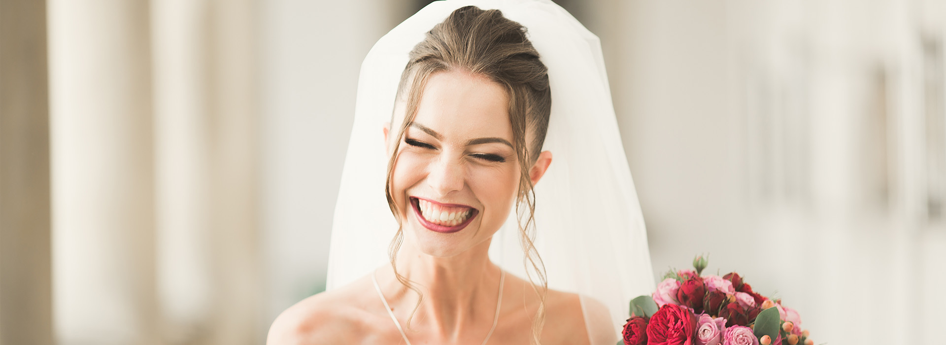 A bride wearing a white wedding dress and veil, smiling happily while holding a bouquet of flowers.