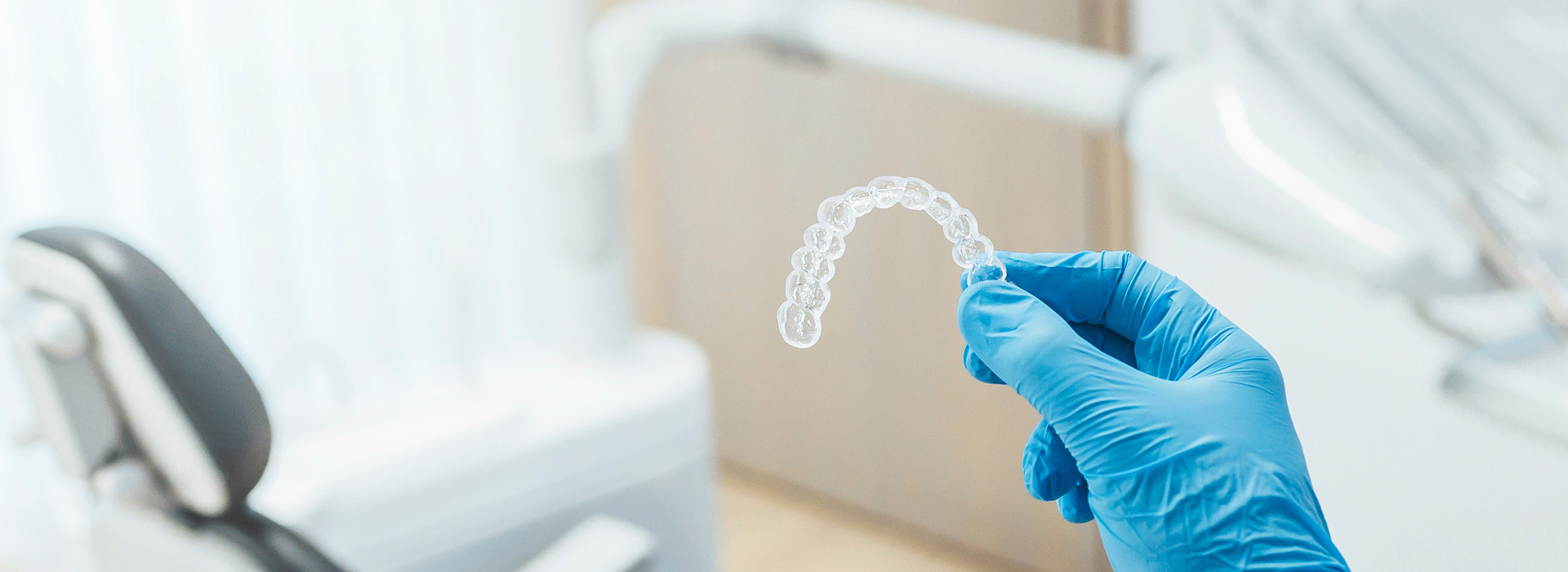 A person wearing blue gloves is holding a transparent dental impression in their hand, which appears to be placed on a white tabletop. The background shows a dental office setting with medical equipment and a brightly lit environment.