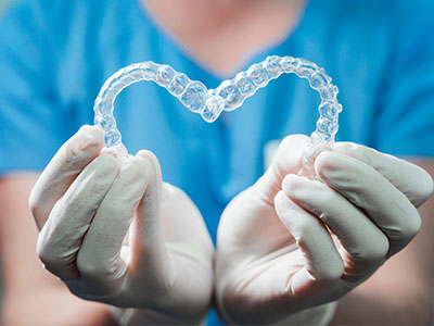 The image features a pair of clear braces with a heart-shaped cutout held by gloved hands against a blue background.