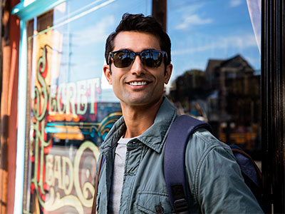 A smiling man in sunglasses, standing outdoors with a backpack, posing for the camera in front of a store window.