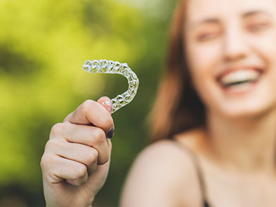 The image features a young woman holding up a clear plastic dental retainer with her left hand, smiling and looking directly at the camera.