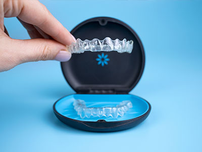 A person s hand holding an open dental impression tray that displays transparent plastic teeth inserts, with a blue background and a watermark of a toothbrush.