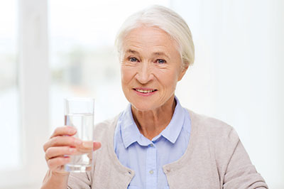 The image features an elderly woman holding a glass of water, smiling at the camera.