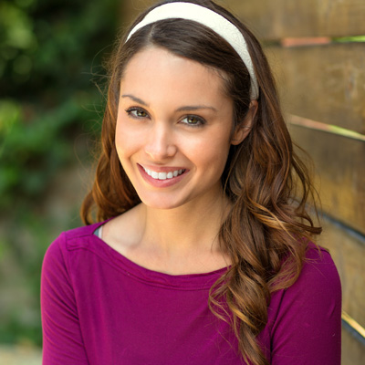 The image shows a woman with long hair, wearing a purple top and a headband, smiling at the camera against a wooden fence background.