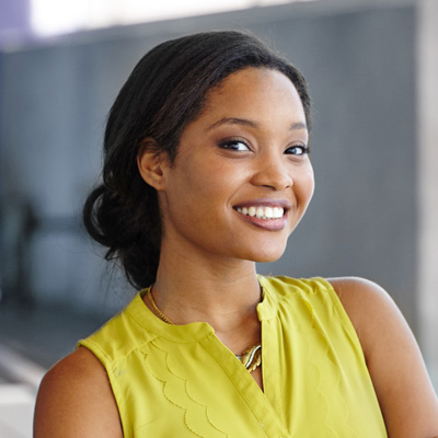 The image features a smiling woman with dark hair, wearing a yellow top and a necklace, standing in front of a building with a large window.