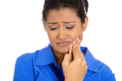 A woman in a blue shirt with her mouth open, looking upwards with a concerned expression, holding what appears to be a toothbrush or dental tool near her teeth.