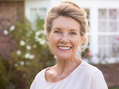 An elderly woman with short hair, wearing a white top, stands in front of a brick house with a garden and a window.