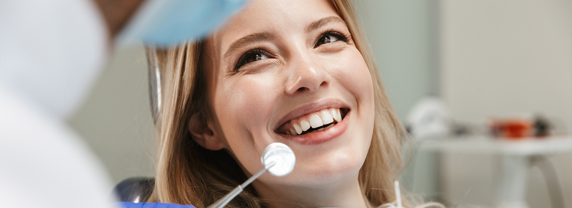 A woman in a dental chair, smiling at the camera, with a dentist performing an examination.