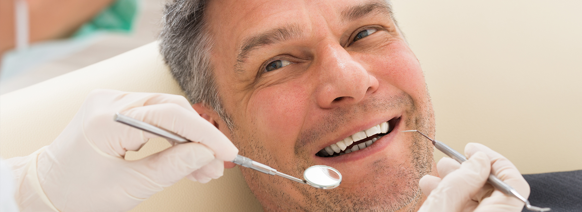 A man is sitting in a dental chair, smiling broadly while receiving oral care from a dentist who is cleaning his teeth.