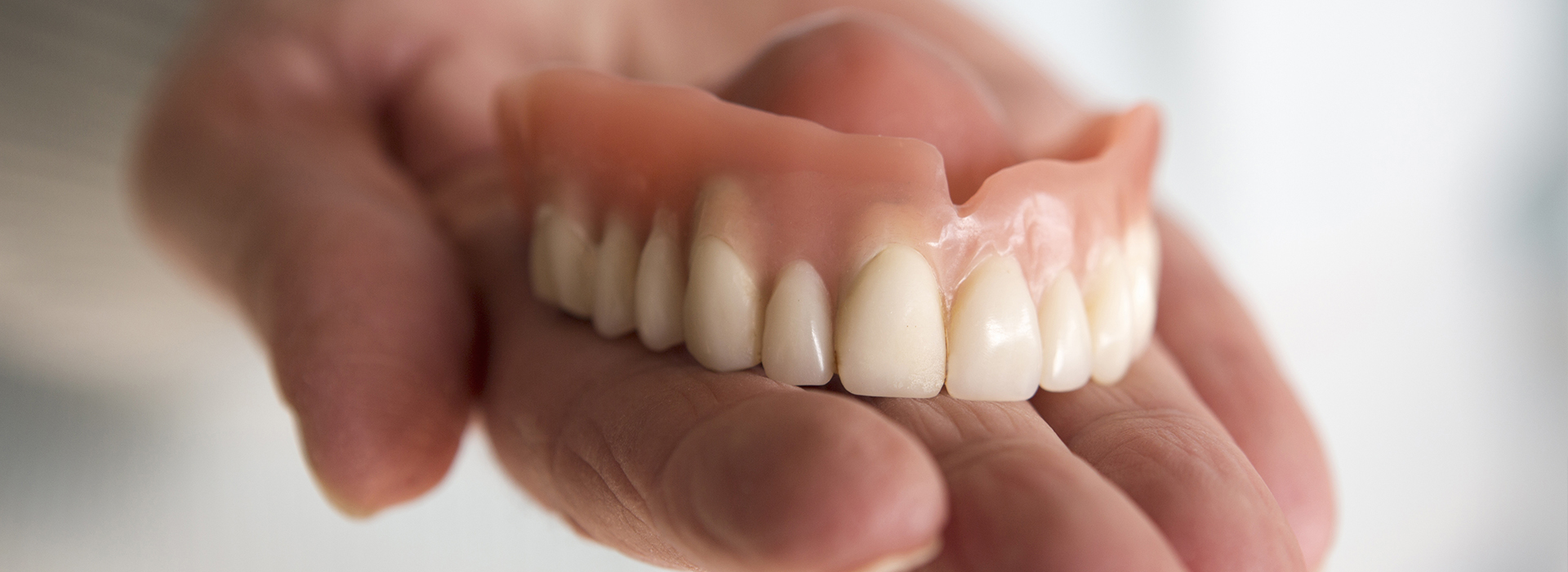 The image shows a person holding up a set of dentures with their left hand, highlighting the upper and lower teeth.