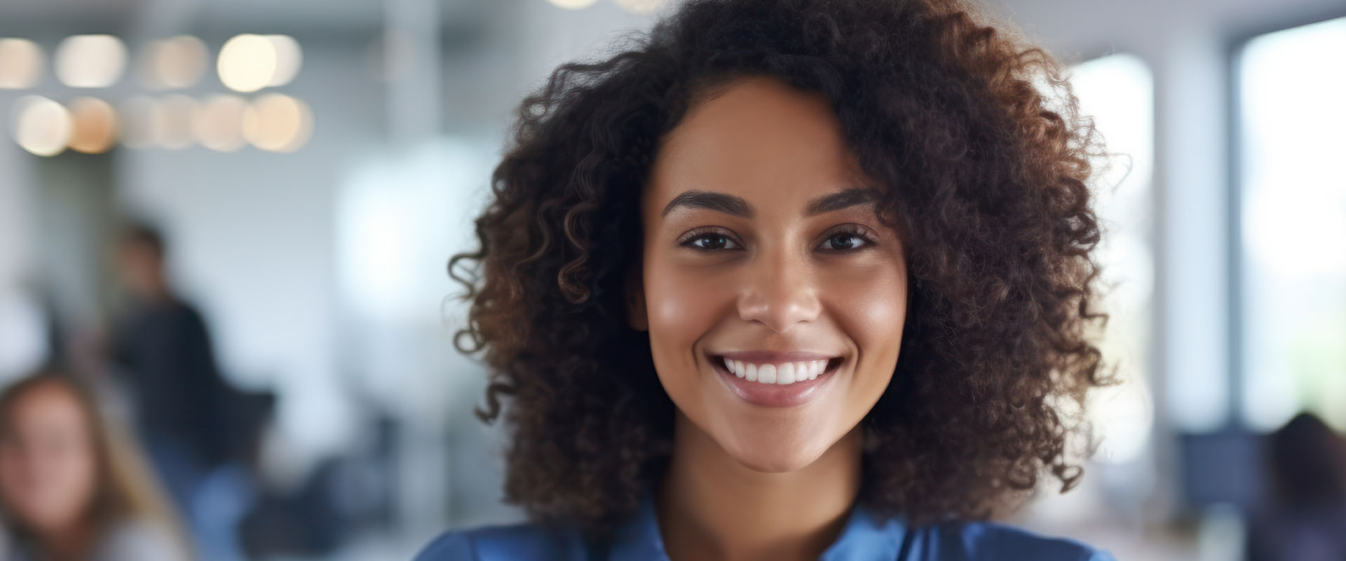 The image is a photograph of a smiling woman with dark hair, wearing a blue top, standing in an indoor office environment.