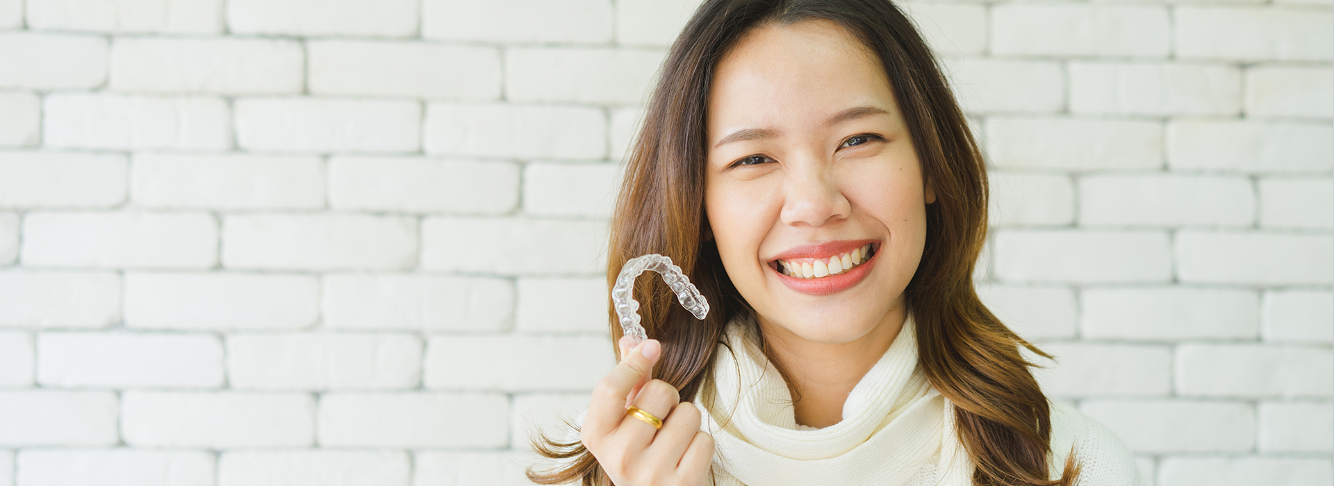 A woman with a radiant smile is holding a ring, standing in front of a brick wall.