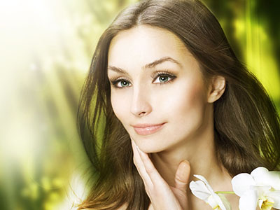 A woman with long hair, wearing makeup and a serene expression, set against a blurred background featuring greenery.