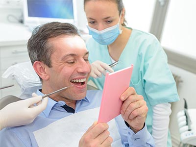 The image shows a man sitting in a dental chair, holding a pink card with a smile, while looking at it. Behind him, a female dental professional is standing with her hands on the chair s armrests, wearing a surgical mask and gloves, and appears to be laughing or smiling as she looks towards the camera. The setting suggests a dental office environment with medical equipment visible in the background.