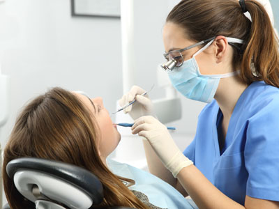 A dental hygienist is assisting a woman in a dental chair, with both wearing masks and working with dental equipment.