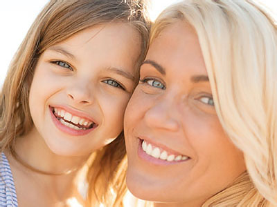 The image shows a woman and a young child, both smiling, with the woman s face partially obscured by the child s head. They appear to be outdoors in bright sunlight, possibly on a beach or similar location.