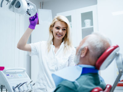 A dental hygienist in a white uniform is assisting an elderly patient with a toothbrush in a dental office.