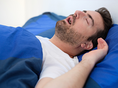 A man in a white t-shirt is lying on his back in bed with his eyes closed, appearing to be asleep.