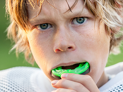 The image shows a young male athlete with blonde hair, wearing a football uniform and holding a green sports mouthguard in his mouth.