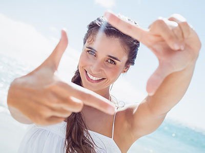 The image features a woman taking a selfie with her hand in front of her face, set against a bright beach backdrop.